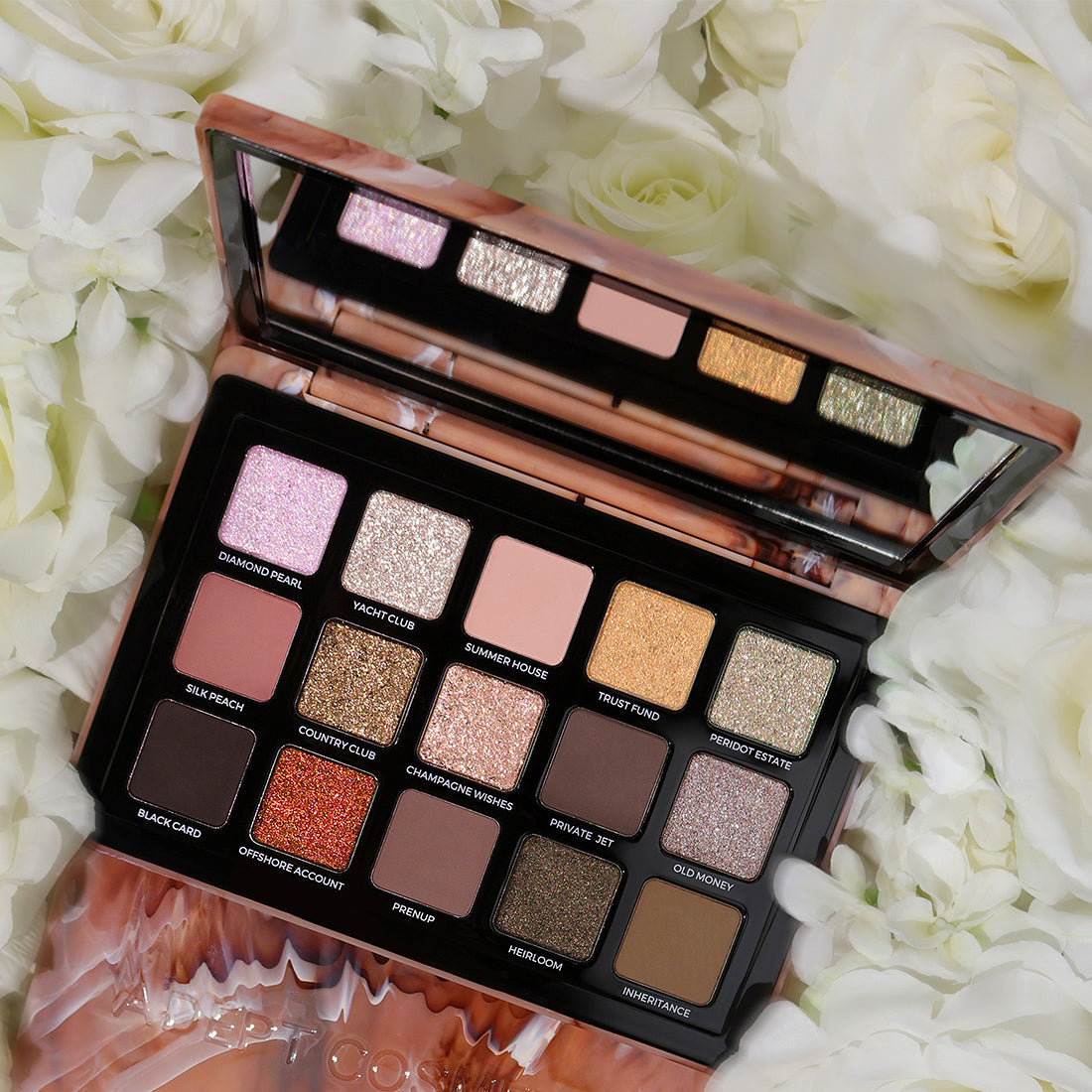 An open 12-shade eyeshadow palette with a rose gold metallic case, surrounded by white roses. The palette contains a mix of matte, shimmer, and glitter shades in pink, gold, brown, and bronze tones, with the brand name 'COSMIC' visible on the lower portion of the case.