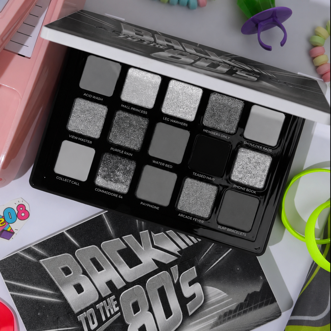 A Back to the 80's themed eyeshadow palette with 12 shades in black, gray, and silver, displayed alongside its matching box and colorful retro-themed props.