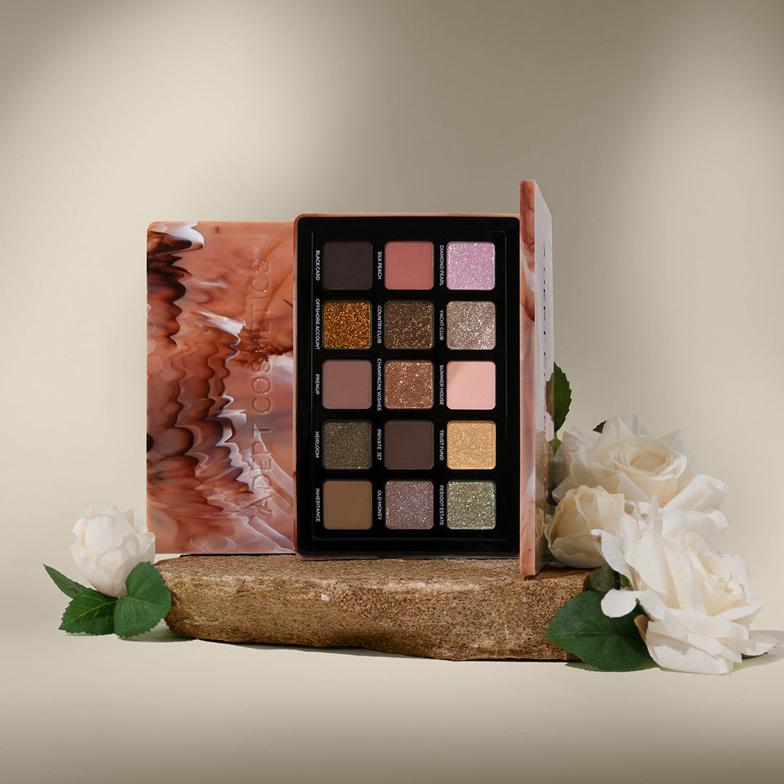 A 15-shade eyeshadow palette displayed open on a rustic wooden block, flanked by a marbled pink and orange case and white roses with green leaves, set against a warm beige gradient background.