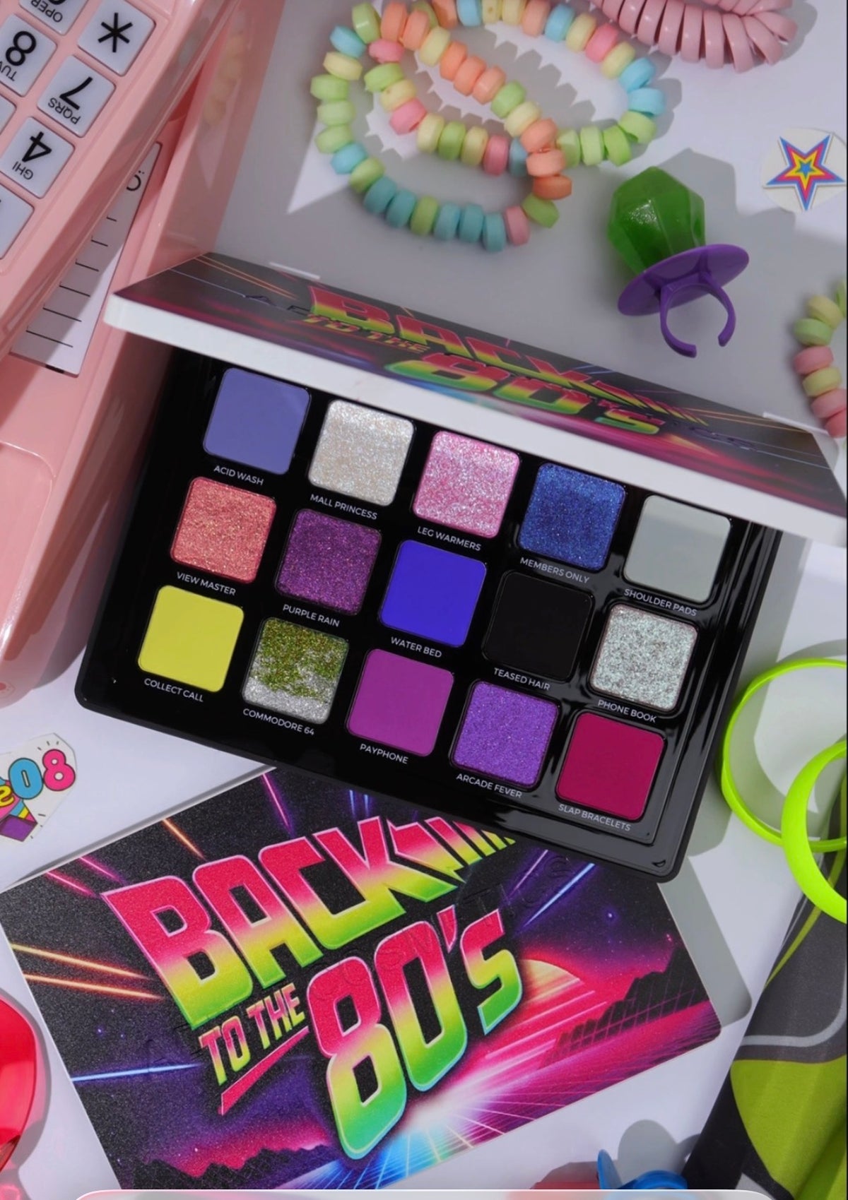 A 15-shade eyeshadow palette themed after 'Back to the 80's' is shown open, surrounded by retro-themed props including a pink toy phone, pastel candy bracelets, and a pacifier on a white background.